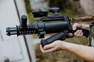 A person holding a tactical weapon with a cylindrical barrel and attached scope, standing outdoors. The weapon is predominantly black with a textured surface and appears to be designed for military or law enforcement use.