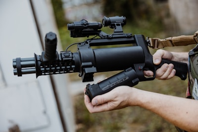 A person holding a tactical weapon with a cylindrical barrel and attached scope, standing outdoors. The weapon is predominantly black with a textured surface and appears to be designed for military or law enforcement use.