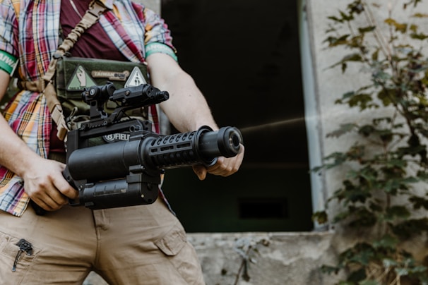 A person in a plaid shirt holds a large black gun while standing in front of an open door to a building. The person is wearing a vest with various patches and equipment. The background features some plants and an outdoor setting.