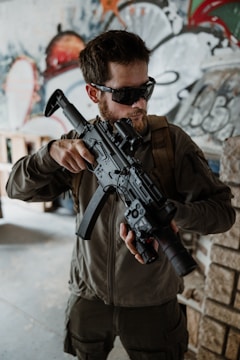A man wearing tactical gear and sunglasses is holding a large camera with a long lens, resembling a weapon. The background features abstract graffiti art in various colors.