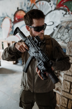 A man wearing tactical gear and sunglasses is holding a large camera with a long lens, resembling a weapon. The background features abstract graffiti art in various colors.