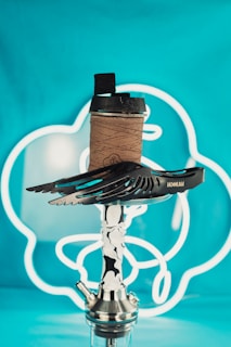A modern-looking hookah with a sleek metal design and a decorative wing-shaped coal holder on top. It is set against a bright turquoise background with white neon lights forming abstract shapes. The hookah has a wooden coal holder with black charcoals on top.