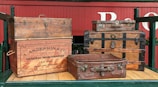 Several vintage and ornate wooden trunks and suitcases are stacked on a wooden platform. The items include a large trunk with a company logo 'A. Anderson & Co General Merchandise' and an establishment date of 1886. The red wagon background and the suitcases give a rustic and historical feel.