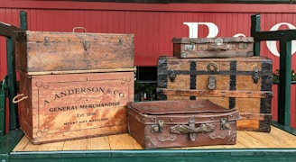 Several vintage and ornate wooden trunks and suitcases are stacked on a wooden platform. The items include a large trunk with a company logo 'A. Anderson & Co General Merchandise' and an establishment date of 1886. The red wagon background and the suitcases give a rustic and historical feel.