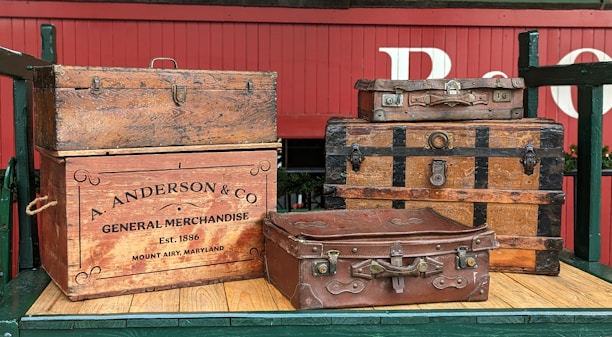 Several vintage and ornate wooden trunks and suitcases are stacked on a wooden platform. The items include a large trunk with a company logo 'A. Anderson & Co General Merchandise' and an establishment date of 1886. The red wagon background and the suitcases give a rustic and historical feel.