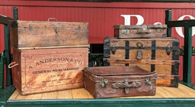 Several vintage and ornate wooden trunks and suitcases are stacked on a wooden platform. The items include a large trunk with a company logo 'A. Anderson & Co General Merchandise' and an establishment date of 1886. The red wagon background and the suitcases give a rustic and historical feel.