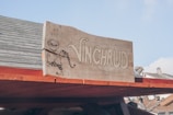 A wooden sign with the word 'VINCHAUD' written on it, accompanied by a simple illustration of a character holding a long bow and a cup. The sign is mounted on the roof of a wooden structure with part of a house visible in the background.