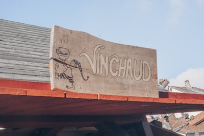 A wooden sign with the word 'VINCHAUD' written on it, accompanied by a simple illustration of a character holding a long bow and a cup. The sign is mounted on the roof of a wooden structure with part of a house visible in the background.