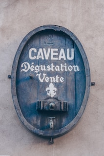 A wooden barrel sign attached to a wall displaying the words 'Caveau Dégustation Vente' in white lettering, featuring a fleur-de-lis symbol underneath.