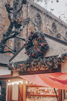A quaint wooden stall is adorned with festive decorations, including a large, ornate wreath featuring a bird motif. String lights twinkle along the garland framing the roof. Below, various Christmas ornaments are on display. Bare tree branches and a stone church facade create a wintry backdrop.