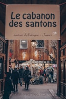 An outdoor market scene with a sign overhead reading 'Le cabanon des santons'. People are walking through a cozy, narrow alleyway adorned with wooden structures and bright string lights. Various booths display goods, and some individuals are holding umbrellas.