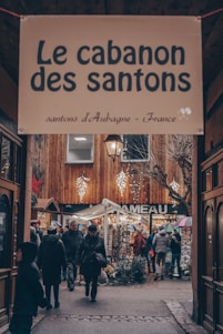 An outdoor market scene with a sign overhead reading 'Le cabanon des santons'. People are walking through a cozy, narrow alleyway adorned with wooden structures and bright string lights. Various booths display goods, and some individuals are holding umbrellas.