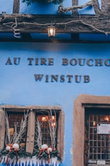 A rustic building facade painted in bright blue with wooden elements. The lettering 'AU TIRE BOUCHON WINSTUB' is prominently displayed. Festive decorations with white branches, red and white ornaments adorn the window sill. A vintage lantern hangs above the sign, casting a warm glow.