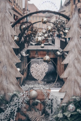 A decorative setup featuring two wooden cutouts shaped like trees, framing a woven heart decoration in the center. There are various Christmas ornaments, including gold and silver baubles, hanging in the background. The scene appears urban, with buildings and a Ferris wheel visible in the distance.
