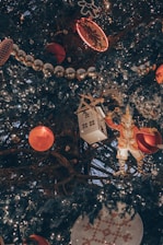 A close-up view of a decorated Christmas tree adorned with various ornaments, including red baubles, a white house ornament with a wooden roof, a figure resembling a toy soldier or nutcracker, strands of shiny silver beads, and a gingerbread-shaped light. The tree is illuminated with numerous string lights, creating a warm and festive atmosphere.
