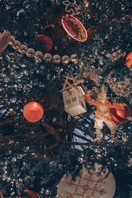 A close-up view of a decorated Christmas tree adorned with various ornaments, including red baubles, a white house ornament with a wooden roof, a figure resembling a toy soldier or nutcracker, strands of shiny silver beads, and a gingerbread-shaped light. The tree is illuminated with numerous string lights, creating a warm and festive atmosphere.