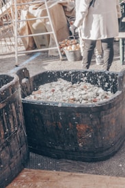 A large wooden barrel filled with a mixture that appears to be fermenting, surrounded by a market or outdoor fair setting. To the right, a person wearing a light-colored coat is visible from the back, standing near containers of onions and other vegetables. A metal ladder and fabric-covered items are in the background.