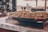 A large metal pan filled with cooked nuts and dried fruits is placed on a table covered with a patterned cloth. The scene appears to be in an outdoor setting, possibly at a food market, with blurred background elements such as people and structures.