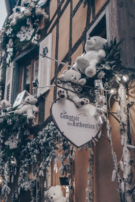 A quaint shop façade adorned with plush white teddy bears and festive decorations. The foreground features a heart-shaped sign with text and decorative branches with ornaments. The background showcases a timber-framed building.
