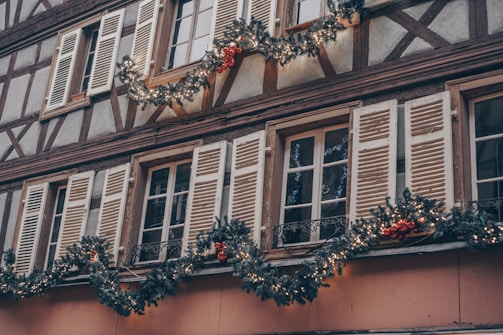 A building facade decorated with festive garlands featuring lights and red ornaments. The walls have a timber frame design, and the windows are adorned with white shutters.