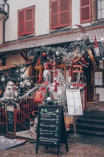 A charming storefront decorated for the holiday season, featuring festive elements such as evergreen garlands with silver baubles, red accents, and whimsical gnome figures. The building has snow-covered window sills, red shutters, and a rustic tiled roof. A chalkboard menu stands on the cobblestone sidewalk, displaying various dishes in elegant handwriting.