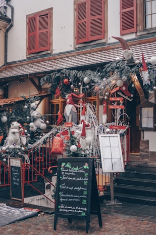A charming storefront decorated for the holiday season, featuring festive elements such as evergreen garlands with silver baubles, red accents, and whimsical gnome figures. The building has snow-covered window sills, red shutters, and a rustic tiled roof. A chalkboard menu stands on the cobblestone sidewalk, displaying various dishes in elegant handwriting.