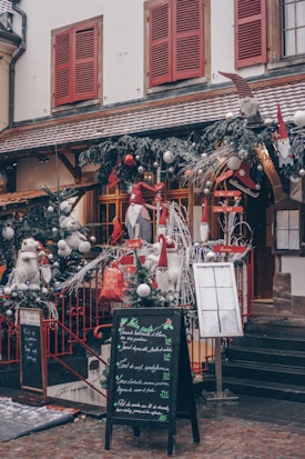 A charming storefront decorated for the holiday season, featuring festive elements such as evergreen garlands with silver baubles, red accents, and whimsical gnome figures. The building has snow-covered window sills, red shutters, and a rustic tiled roof. A chalkboard menu stands on the cobblestone sidewalk, displaying various dishes in elegant handwriting.