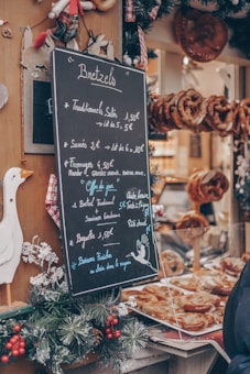 A chalkboard menu listing prices of various pretzels and other items is displayed outside a bakery. The menu is surrounded by festive holiday decorations such as pine branches and red berries. Behind the glass, freshly baked pretzels are visible, adding to the inviting atmosphere.
