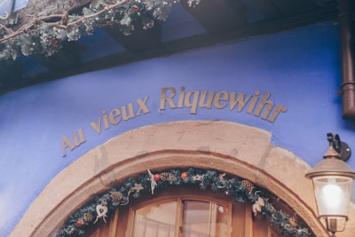 An exterior wall painted in vivid blue with the sign 'Au vieux Riquewihr' in gold lettering above a wooden door. Holiday decorations featuring pine branches, ornaments, and pine cones adorn the entrance. A vintage-style lamp is mounted beside the door.