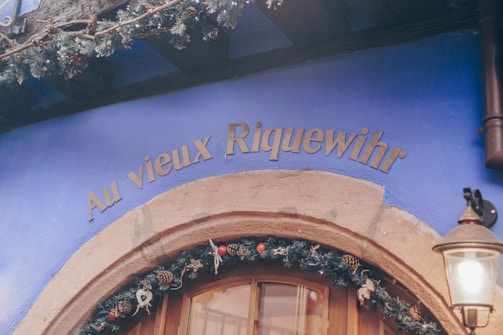An exterior wall painted in vivid blue with the sign 'Au vieux Riquewihr' in gold lettering above a wooden door. Holiday decorations featuring pine branches, ornaments, and pine cones adorn the entrance. A vintage-style lamp is mounted beside the door.