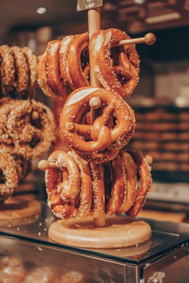 A wooden stand displays several freshly baked pretzels adorned with coarse salt, arranged in a bakery setting. The pretzels have a golden brown color, suggesting they are deliciously crispy outside while soft inside. In the background, blurred images of additional bakery items are visible.