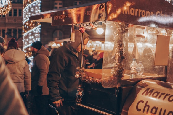 A crowded outdoor market scene during the evening, featuring a person selling items from a decorated stall adorned with tinsel. The stall displays a sign with words partially visible like 'Marrons Chaud.' People are dressed warmly, suggesting a cold atmosphere, with festive lights illuminating the area, enhancing the market's cozy ambiance.