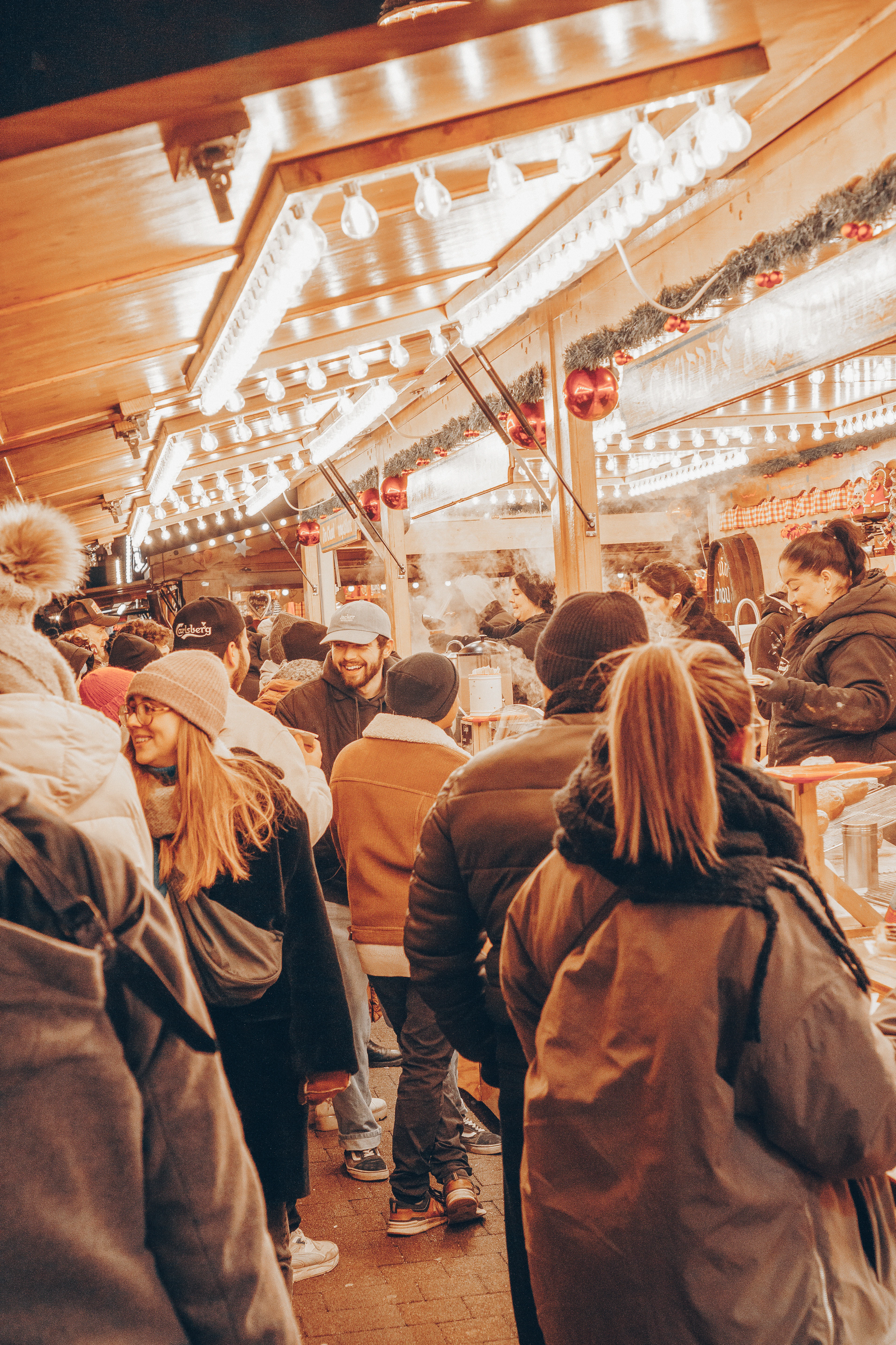 A crowded, lantern-lit Christmas market stall glows with string lights and evergreen garlands at dusk.
