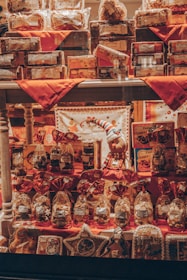 A shop window display filled with various packages and gift items, including cookies, biscuits, and holiday-themed decorations. The arrangement features red accents with decorative bags and boxes, and a small stuffed figure wearing festive attire is centrally placed.
