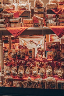 A shop window display filled with various packages and gift items, including cookies, biscuits, and holiday-themed decorations. The arrangement features red accents with decorative bags and boxes, and a small stuffed figure wearing festive attire is centrally placed.