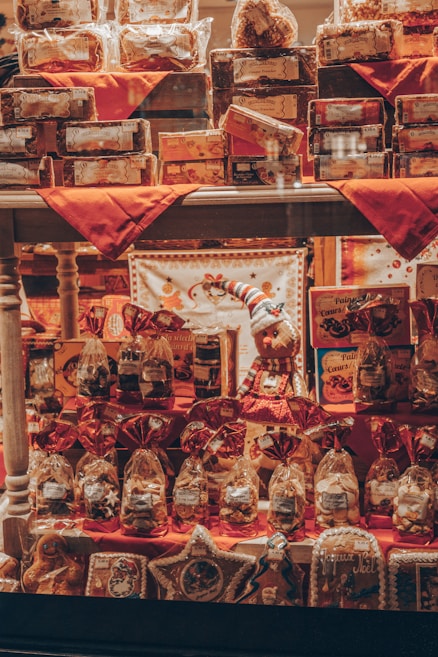 A shop window display filled with various packages and gift items, including cookies, biscuits, and holiday-themed decorations. The arrangement features red accents with decorative bags and boxes, and a small stuffed figure wearing festive attire is centrally placed.