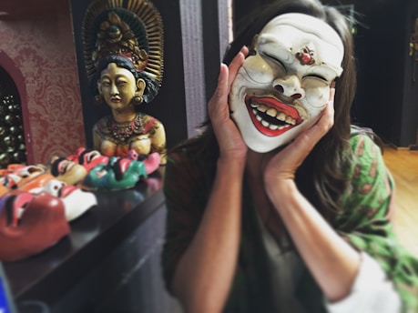 A person is holding a white traditional mask with an exaggerated laughing expression, covering their face. In the background, there is a shelf with multiple colorful masks and a detailed statue of a figure with an ornate headdress and jewelry. The setting appears to be indoors with warm lighting.