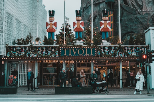 The entrance of an amusement park, decorated with festive elements and large toy soldier statues on top. People are gathered at the entrance, some with children and a stroller. The facade is adorned with lights, garlands, and Christmas decorations. The word 'TIVOLI' is prominently displayed above the entrance.