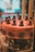 A beautifully crafted chocolate cake with a smooth, glossy surface adorned with small, intricate chocolate figurines around the top edge. The cake is decorated with round, embossed chocolate discs along the side, and it sits on a dark, reflective cake stand inside a display case.