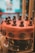 A beautifully crafted chocolate cake with a smooth, glossy surface adorned with small, intricate chocolate figurines around the top edge. The cake is decorated with round, embossed chocolate discs along the side, and it sits on a dark, reflective cake stand inside a display case.
