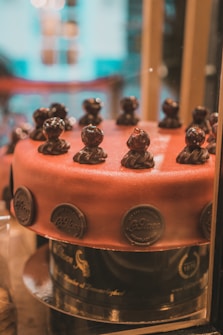 A beautifully crafted chocolate cake with a smooth, glossy surface adorned with small, intricate chocolate figurines around the top edge. The cake is decorated with round, embossed chocolate discs along the side, and it sits on a dark, reflective cake stand inside a display case.