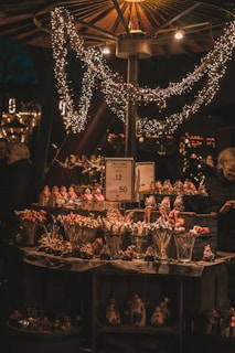 A cozy market stall illuminated by warm lights and decorated with sparkling string lights. The stall is filled with festive items, including small figurines and lollipops shaped like different objects, suggesting a holiday theme. A price sign is visible, indicating the sale of these items, and a person stands nearby, partially in view.