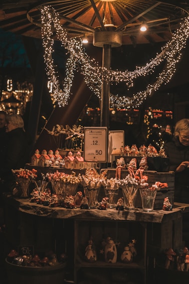 A cozy market stall illuminated by warm lights and decorated with sparkling string lights. The stall is filled with festive items, including small figurines and lollipops shaped like different objects, suggesting a holiday theme. A price sign is visible, indicating the sale of these items, and a person stands nearby, partially in view.