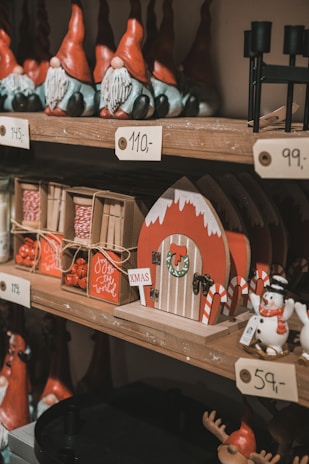 A wooden shelf displays a festive assortment of Christmas decorations, including figurines of gnomes with red hats and long white beards, a set of boxes wrapped in twine, a snowman, and a decorative red house adorned with a wreath and candy canes. Small placards with prices in a non-English currency are visible.