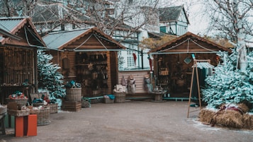 A quaint outdoor market scene featuring wooden stalls adorned with string lights. The stalls contain various items for sale, including baskets, textiles, and other handcrafted goods. Surrounding the stalls are small, snow-dusted evergreen trees and bare birch trees, adding a festive touch. The atmosphere suggests a holiday market set in a rustic, winter setting.