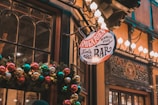 A warmly lit, festive façade of a bar featuring a decorative garland adorned with colorful Christmas baubles. The vintage style sign hanging above, with ornate script, contributes to the cozy, welcoming atmosphere. Illuminated by soft, glowing lights, the doorway is surrounded by dark wood and intricate details.