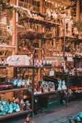 A market stall is adorned with rows of Christmas-themed decorations, including small houses, santa figurines, reindeer, and gnomes. The stall is well-lit with warm, festive lighting and features shelves filled with these items. Some wreaths are hanging, adding to the holiday ambiance.