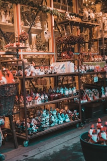 A variety of Christmas-themed figurines and decorations are displayed on metal shelves. The shelves are adorned with small Santa figures, decorative houses, snowmen, wreaths, and miniature Christmas trees. Warm lighting enhances the festive atmosphere, and the background is filled with garlands and more holiday decor.