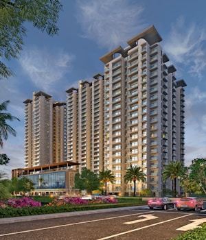 A modern high-rise building complex dominates the skyline, featuring sleek architecture and numerous balconies. Surrounded by landscaped greenery, including palm trees and vibrant flowers, the scene is set against a clear sky at dusk. Cars are visible on the road in the foreground, adding a sense of urban activity.