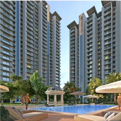 Tall modern residential buildings with numerous windows rise against a clear sky. In the foreground, a swimming pool is surrounded by lounge chairs and umbrellas. A domed pavilion and lush green trees add to the serene setting. A person stands near the pool area.